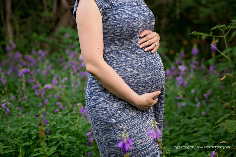 A pregnant Chinese woman hugs her belly while surrounded by purple wildflowers during maternity photos in Edmonton