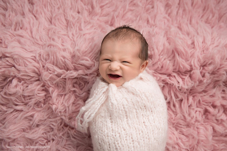 A newborn baby girl smiles as she lays on a pink rug wrapped in a pale pink scarf
