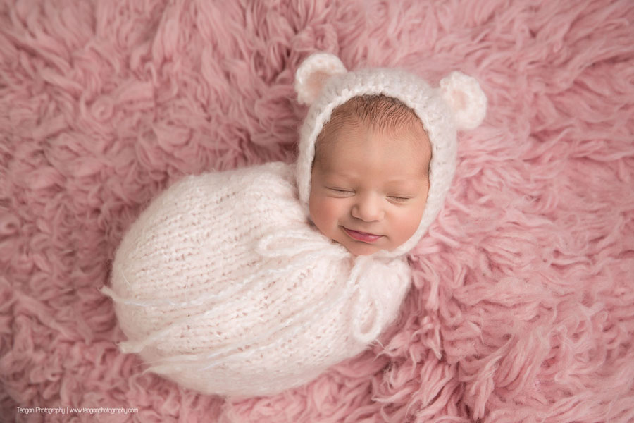 An Edmonton newborn baby sleeps on a pink rug in a pink scarf while wearing a knit pink bear hat