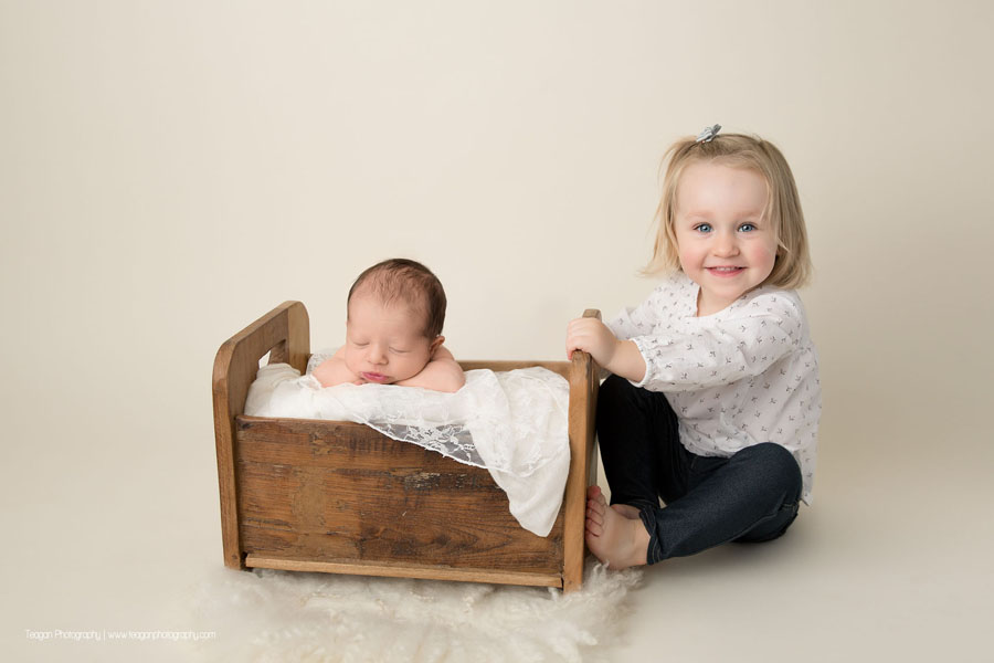 A big sister poses on the floor with her newborn baby who is sleeping in a wooden bed
