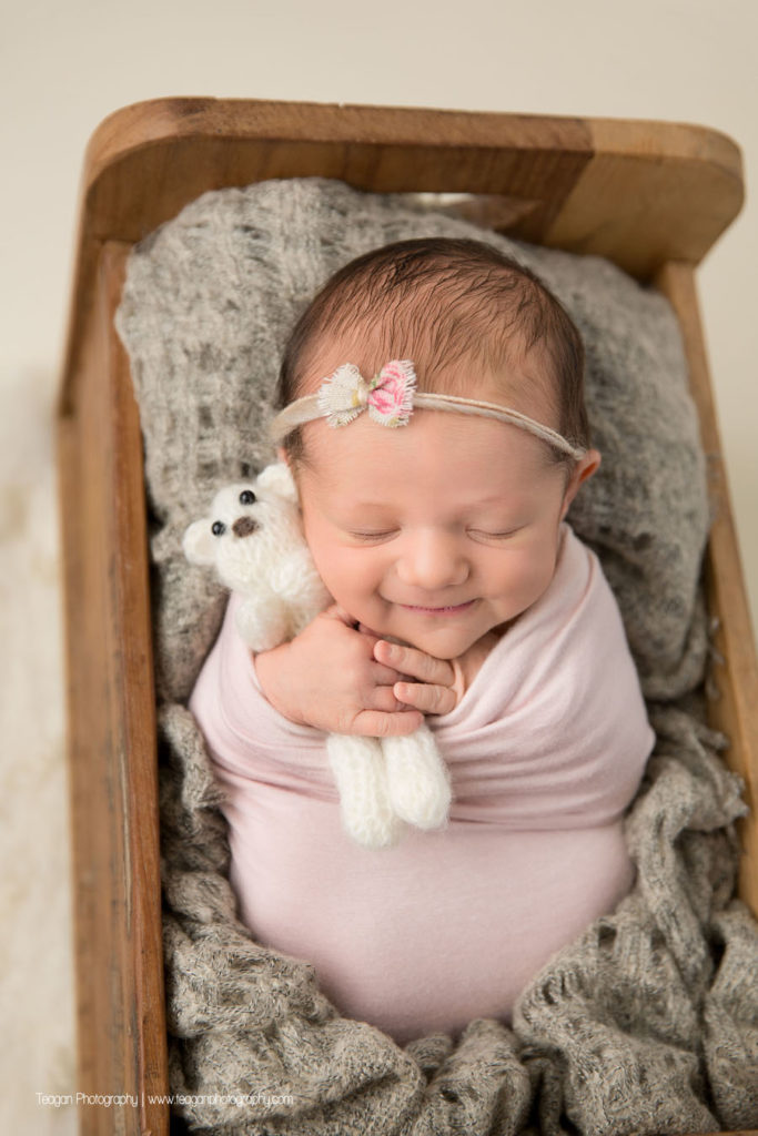 A baby girl is wrapped in pale pink and asleep in a wooden bed while hugging a small white teddy bear