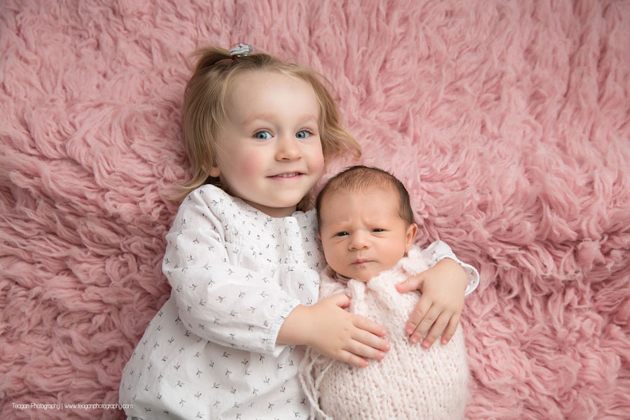 A blonde toddler hugs her newborn baby sister