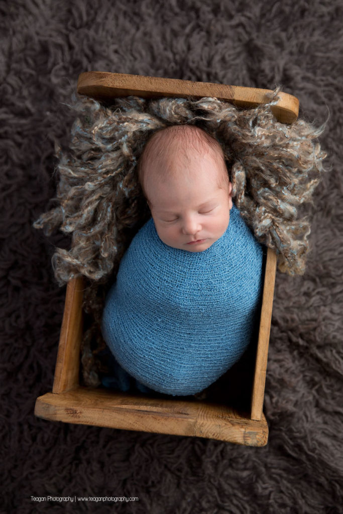 Wrapped in a blue scarf is a newborn baby boy asleep in a wooden bed