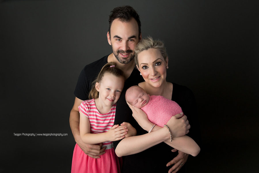 A family wearing pink and black pose together iwith their newborn baby girl during an Edmonton newborn photography session.