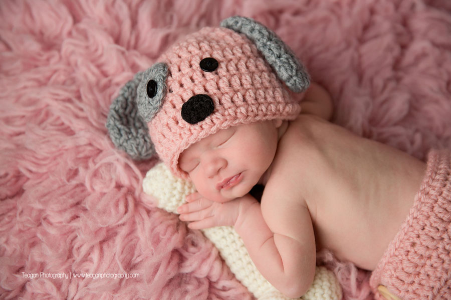 A baby girl is wearing a pink knit puppy bonnet while holding a knit dog bone