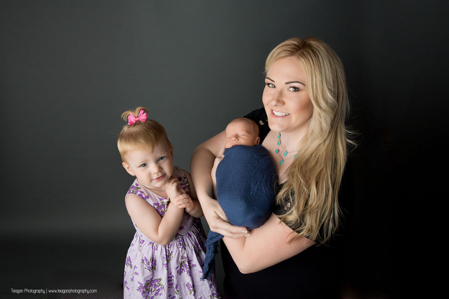 A mom with long blonde hair hold her newborn son while her daughter stands nearby