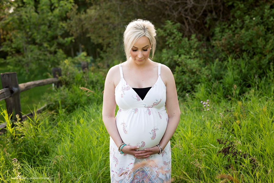 A blonde haired woman wearing a white maternity sun dress hugs her pregnant belly