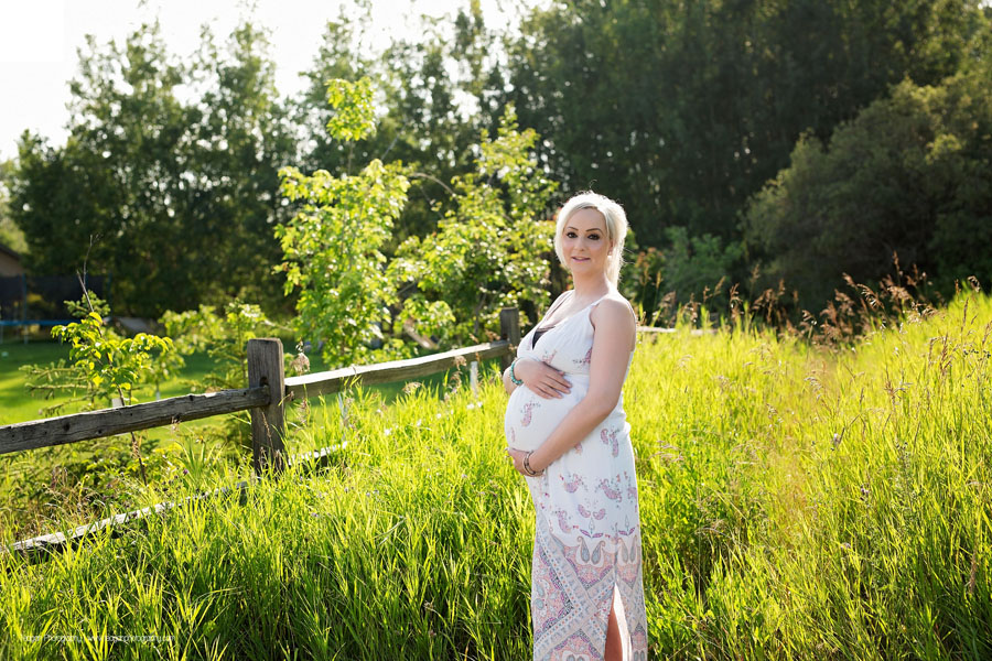 A blonde haired woman wearing a white maternity sun dress hugs her pregnant belly in long field grasses