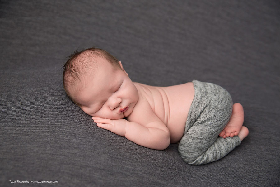 An infant boy sleeps on a grey blanket wearing tiny grey pants