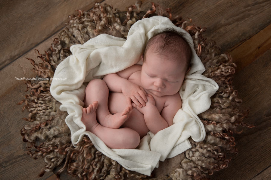 A newborn boy sleeps tucked in a cream colour blanket on a hardwood floor