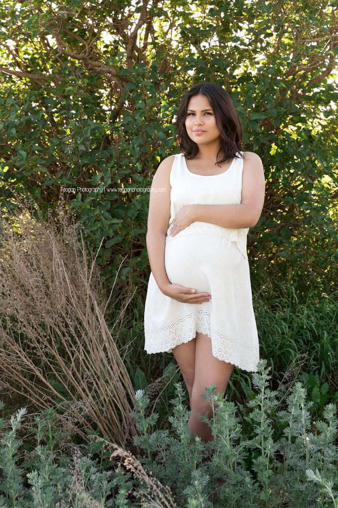 Wearing a white maternity dress is a Native American woman standing amongst sagebrush during an Edmonton maternity photoshoot