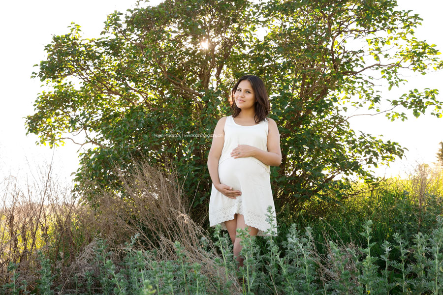 Wearing a white maternity dress is a Native American woman standing amongst sagebrush during an Edmonton maternity photoshoot