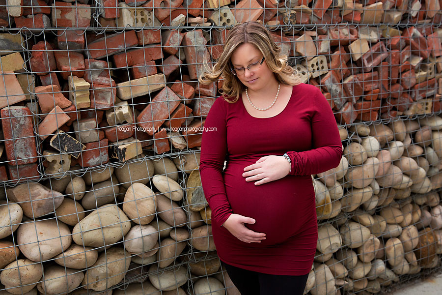 Wearing a burgundy coloured dress is a women hugging her belly during an Edmonton maternity photo shoot