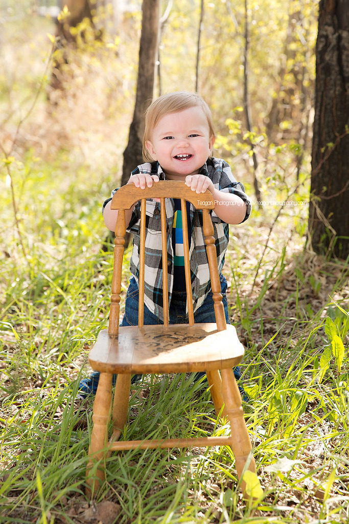 A one year old boy in blue jean overalls poses  with a little chair in the new grass during a Spring photo shoot in Edmonton