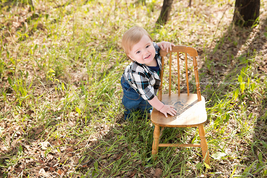 A one year old boy in blue jean overalls poses  with a little chair in the new grass during a Spring photo shoot in Edmonton