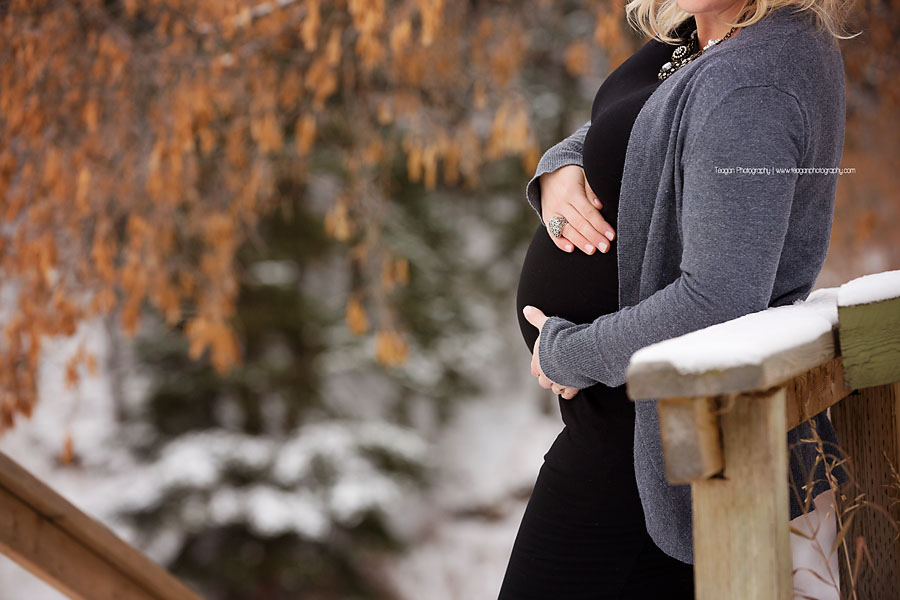 A blonde woman with dark rimmed glasses poses in front of dry orange leaves during a winter maternity session