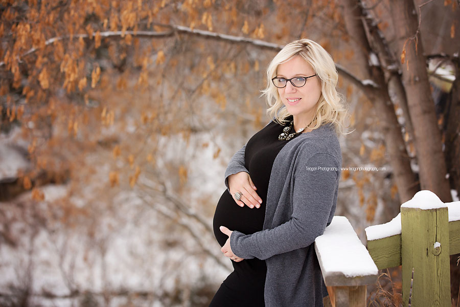 A blonde woman with dark rimmed glasses poses in front of dry orange leaves during a winter maternity session
