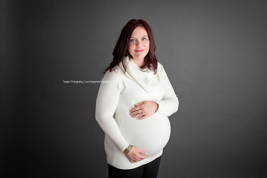 A burgundy haired mother wearing a white sweater poses for maternity photos at a top Edmonton studio