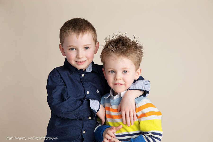 Two blonde preschool aged brothers stand together in front of a beige wall