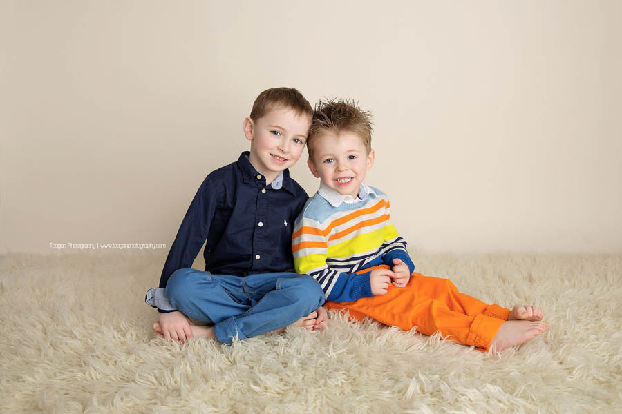 Two blonde preschool aged brothers stand together in front of a beige wall