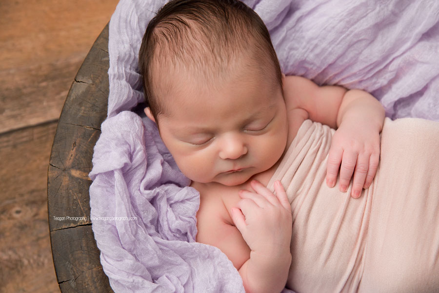 wearing a lavender bear bonnet is a newborn baby girl sleeping on a pale pink blanket 