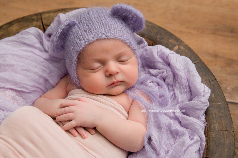wearing a lavender bear bonnet is a newborn baby girl sleeping on a pale pink blanket 