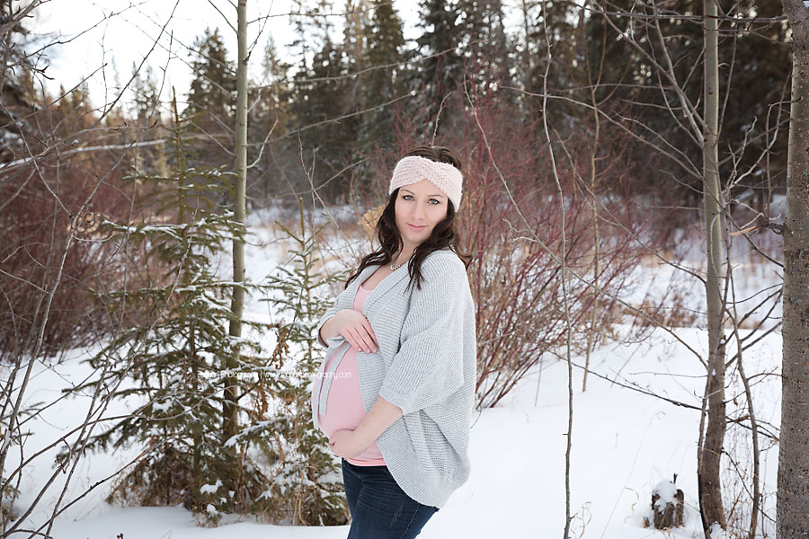 Wearing tones of pink and grey is an Edmonton woman hugging her pregnant belly in the snowy forests of Edmonton
