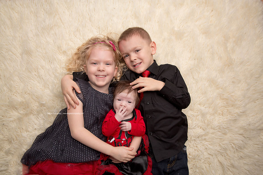 Twin children wearing red and black Christmas clothing hold their newborn sister during an Edmonton family photo shoot in the studio