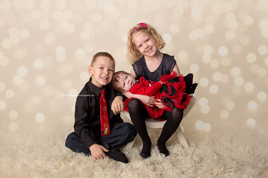 Twin children wearing red and black Christmas clothing hold their newborn sister during an Edmonton family photo shoot in the studio