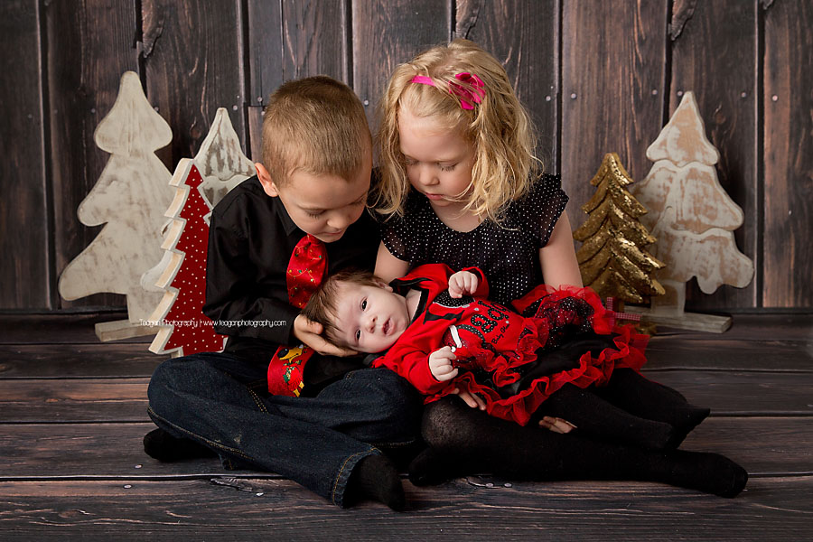 Blonde twin preschool aged children look at their new sister while sitting in front of a Chrismtas themed backdrop in Edmonton