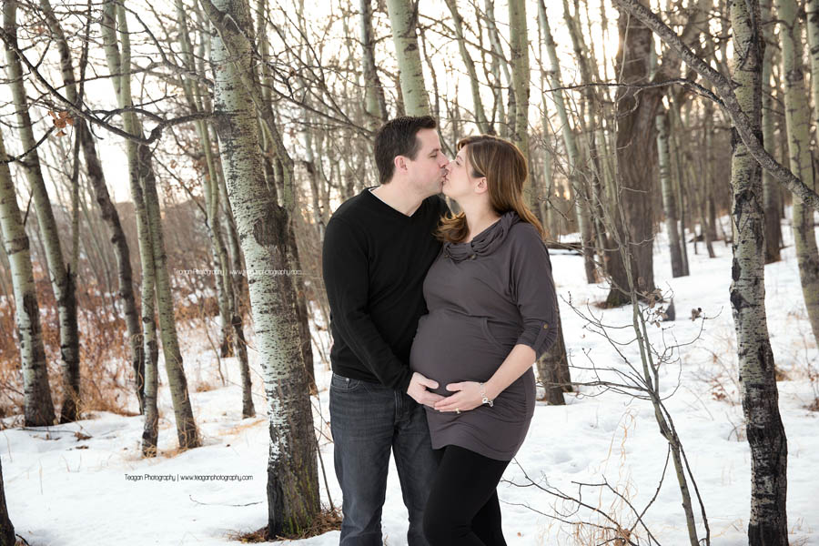A pregnant couple kisses each other in a west Edmonton forest during their winter maternity family photography session