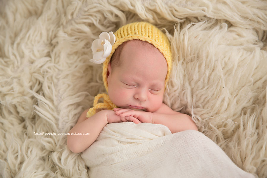 A newborn baby girl wears a butter yellow bonnet while sleeping on a cream coloured rug