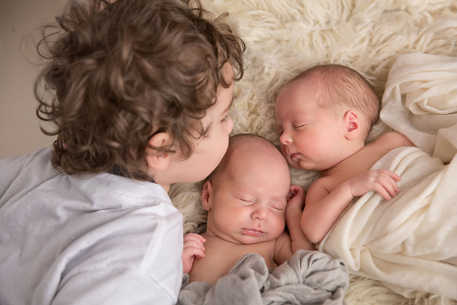 A big brother with brown curly hair kisses his newborn brother and sister