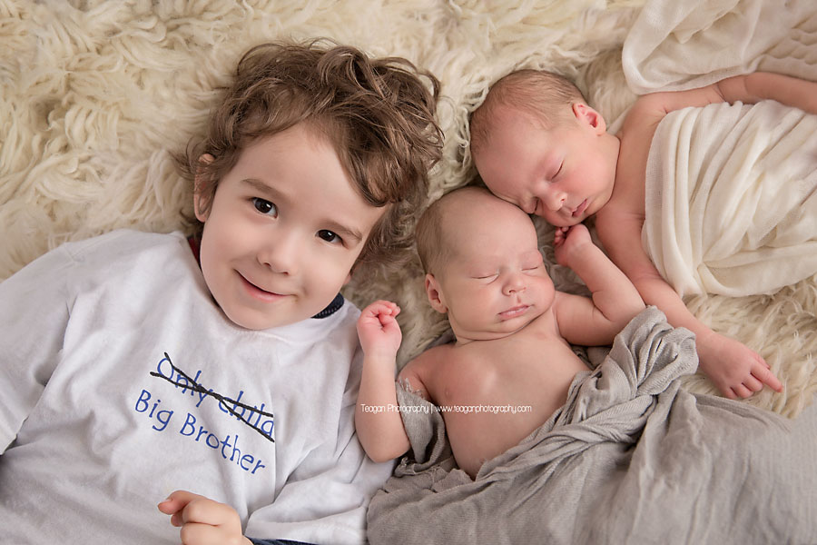 A big brother with brown curly hair kisses his newborn brother and sister