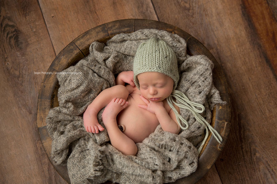 A little baby boy is curled up in a grey blanket and wearing a green knit hat