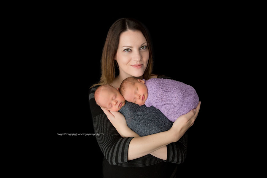 Standing in front of a black backdrop is an Edmonton mother holding her twin newborn babies