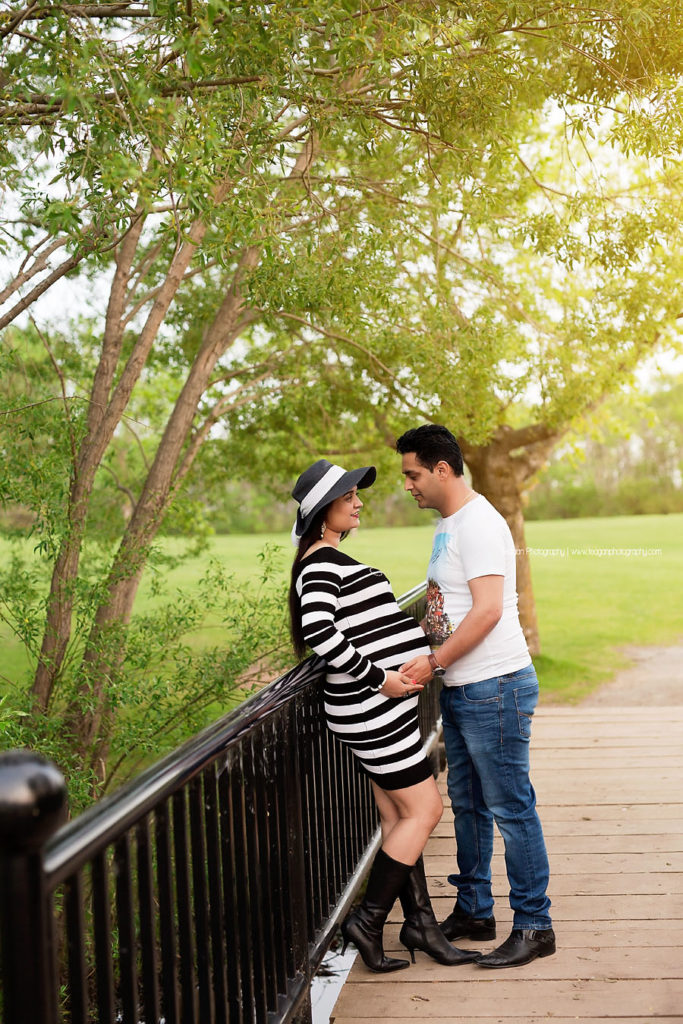 Wearing a white and black striped maternity dress is an hindu woman holding her black hat on a black metal bridge.