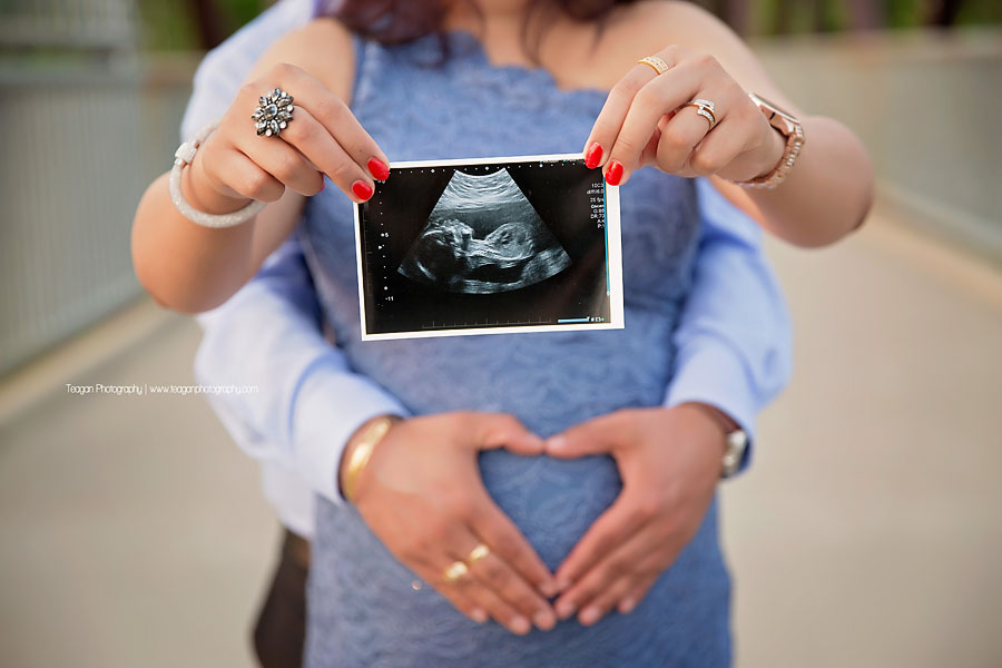 A mother holds up the sonograph photo pf her unborn baby while dad's hands hug her belly.