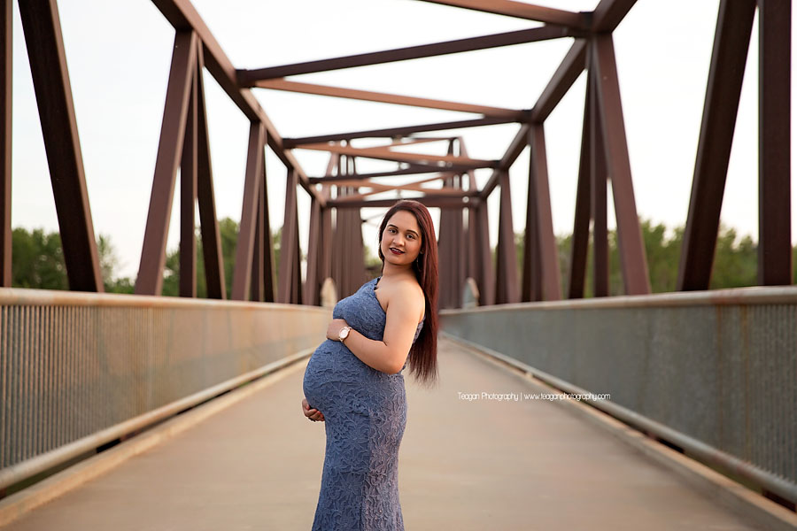 A Hindo woman wearing a perwinkle blue maternity dress poses on the metal bridge to Hawrelak Park for maternity photos