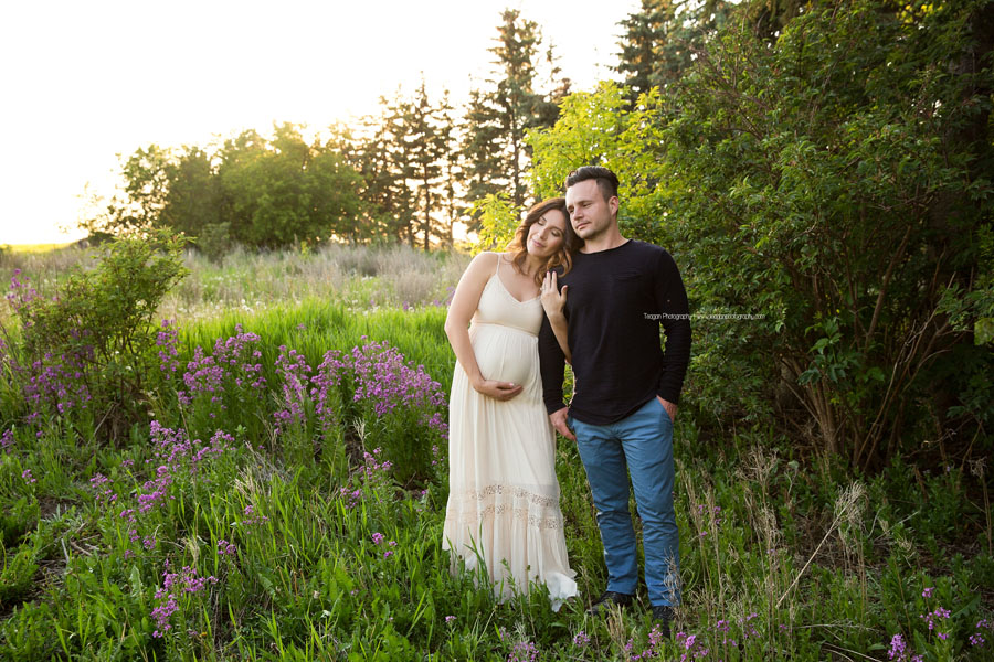 A couple expecting their first baby cuddle together in St Albert's Red Willow Park in the grasses near the wooden train trestle bride