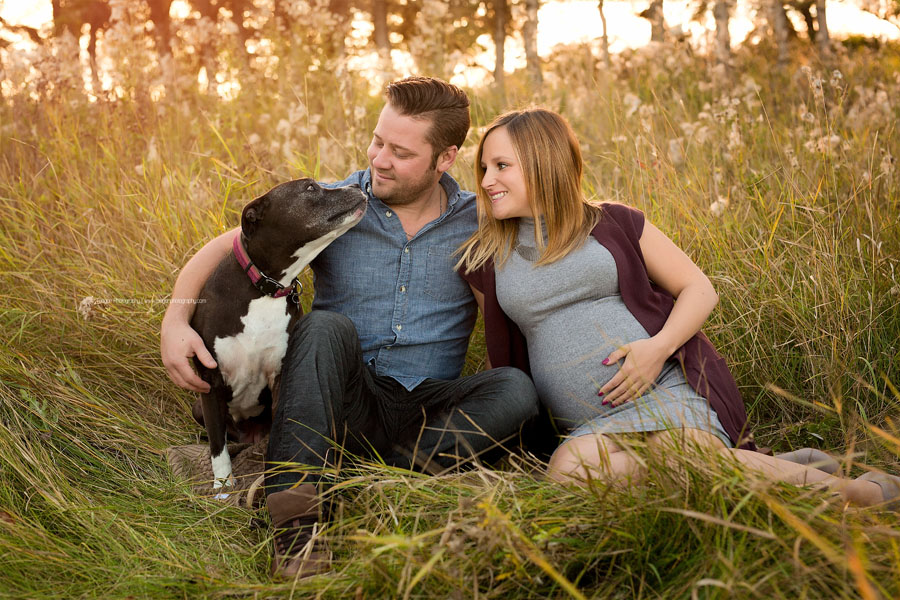 An expectant couple snuggles their dog in the long dry grass during maternity photos.