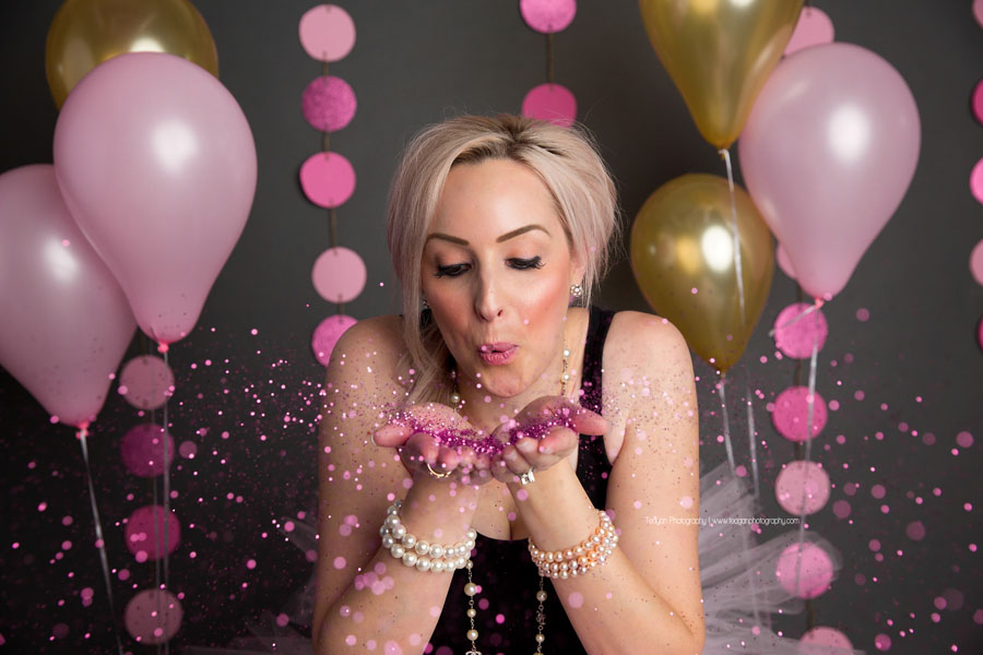 A blonde woman blows pink glitter during her adult cake smash photos in Edmonton