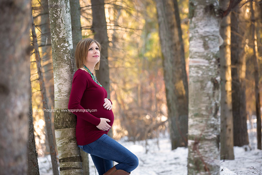 A pregnant woman wearing a red gown poses in a winter snow covered forest in Edmonton