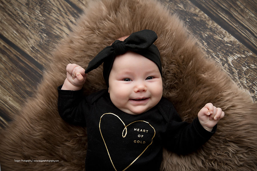 A happy smiling baby in a black dress lays on her back on a brown rug