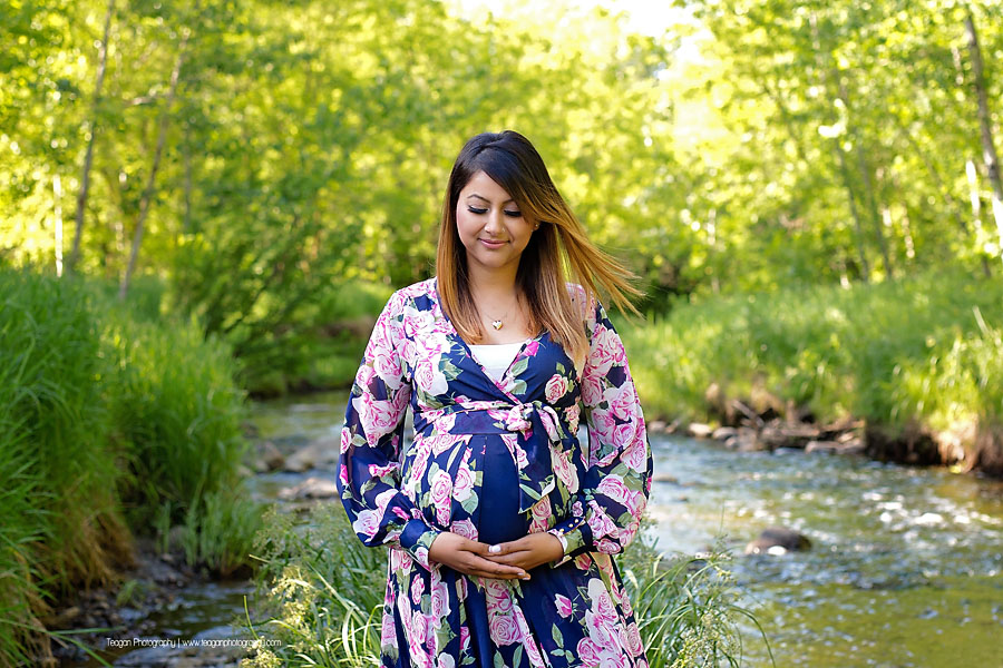 An East Indian mother in a floral gown stands in the middle of a stream in Edmonton during a summer maternity photos. 