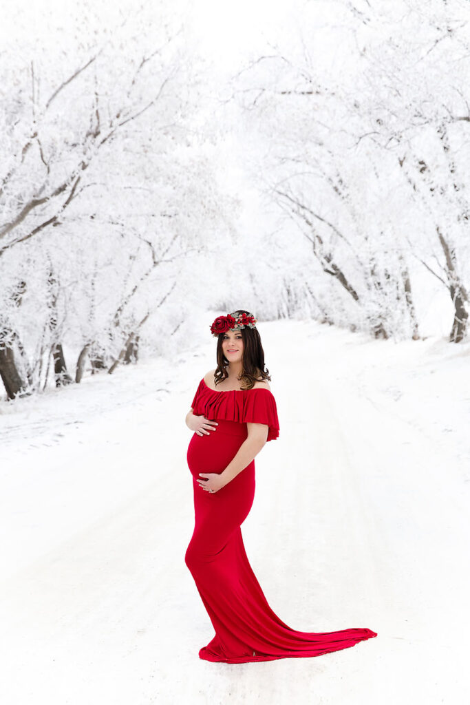 Pregnant mother in a long red maternity dress standing in front of snow-covered trees in Edmonton