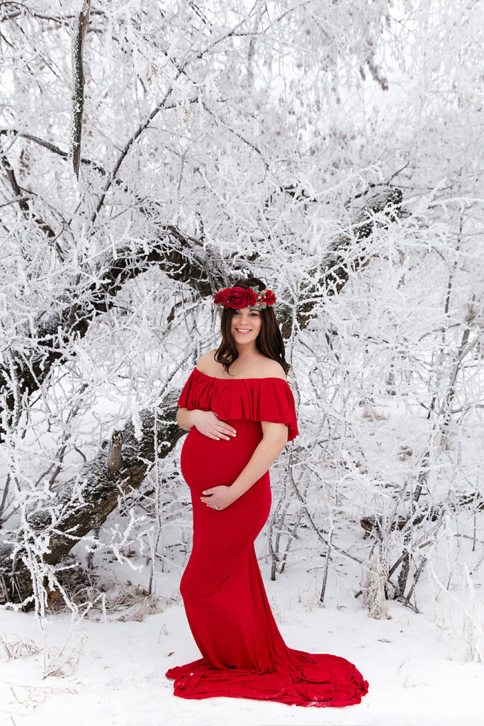 Pregnant mother in a long red maternity dress standing in front of snow-covered trees in Edmonton