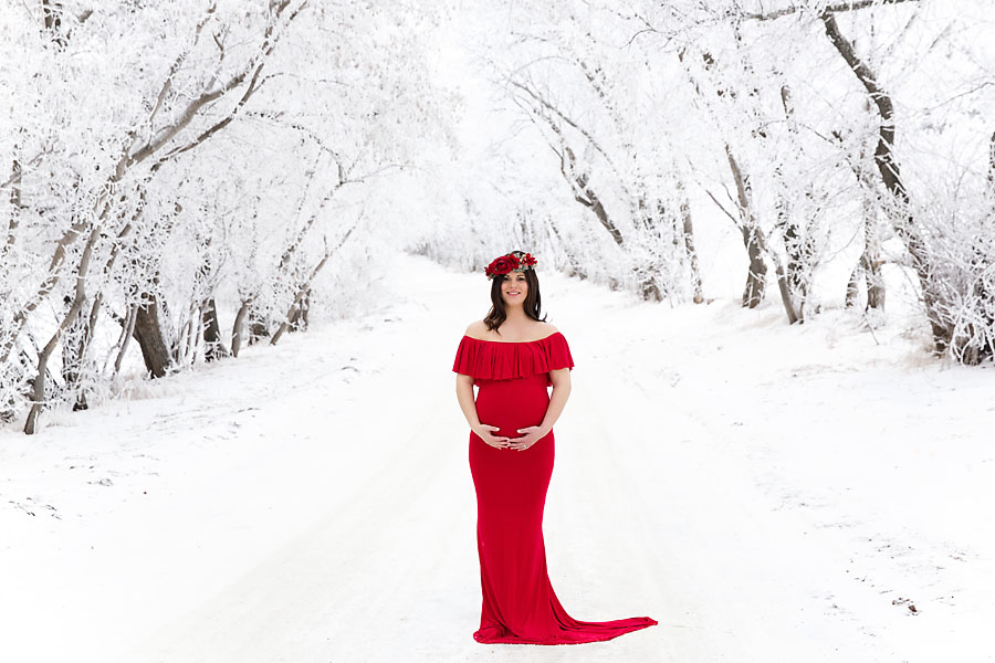 Pregnant mother in a long red maternity dress standing in front of snow-covered trees in Edmonton