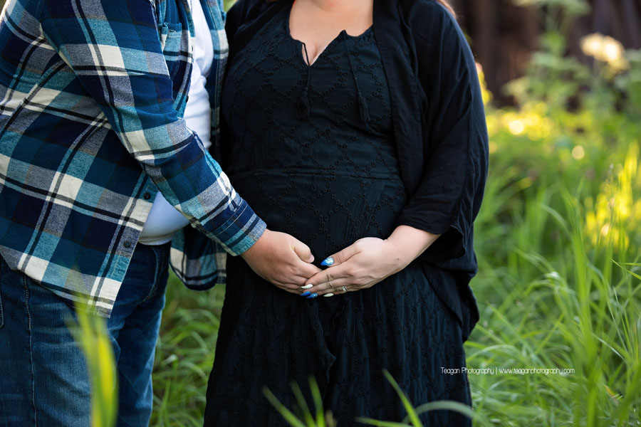 A couple expecting their first baby cuddle together in St Albert's Red Willow Park in the grasses near the  wooden train trestle bride