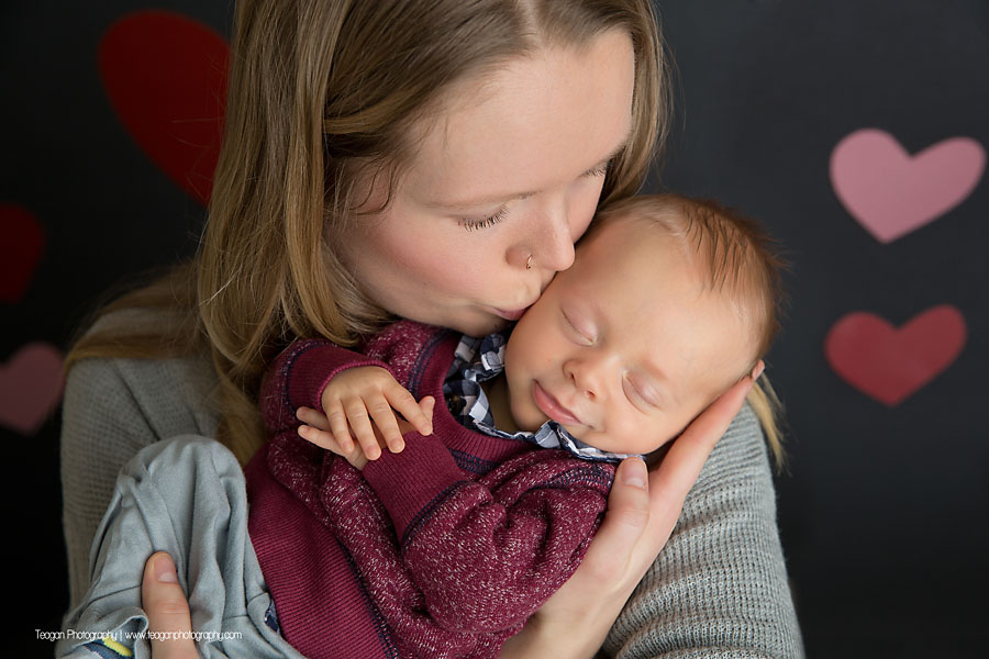 A blond haired mother snuggles with her sleeping  baby in front of a grey wall covered in hearts